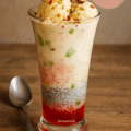 A tall glass of Royal Falooda dessert layered with rose syrup, basil seeds, sev, sweetened milk, topped with vanilla ice cream, nuts, and tutti frutti.