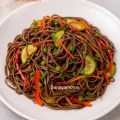 A bowl of colorful vegetable soba noodles tossed in soy-sesame sauce with bell peppers, carrots, and zucchini served on a rustic wooden background.